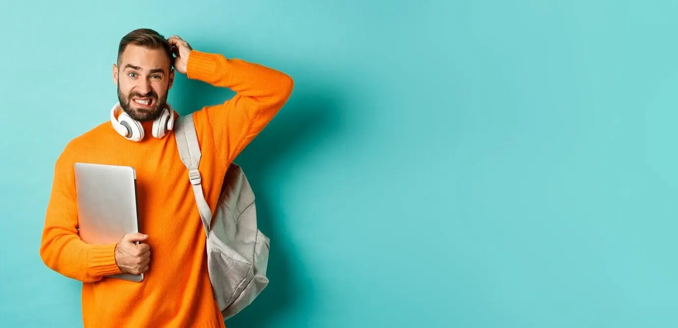 Ein männlicher Student wirkt verwirrt, schaut ratlos, kratzt sich am Kopf und hält Laptop und Rucksack, während er steht.