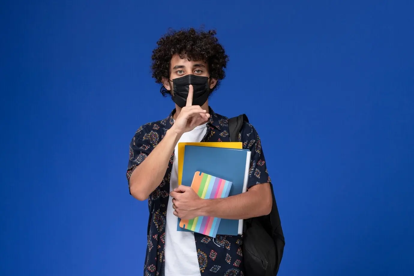 Vorderansicht eines jungen männlichen Studenten mit Rucksack, der eine schwarze Maske trägt und Unterlagen sowie ein Heft hält, vor blauem Hintergrund.